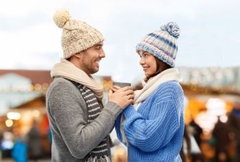 people, winter holidays and love concept - happy romantic couple in knitted hats and scarves holding one cup over christmas market in old town of tallinn city background. happy couple holding one cup at christmas market
