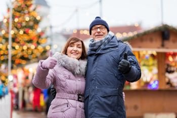 winter holidays and people concept - happy senior couple showing thumbs up at christmas market on town hall square in tallinn, estonia. senior couple shows thumbs up at christmas market