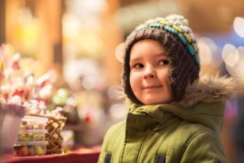holidays, childhood and people concept - happy little boy at christmas market candy shop in winter evening. happy little boy at christmas market shop
