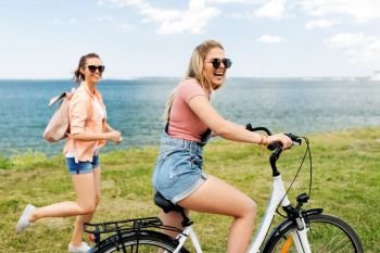 leisure and friendship concept - happy smiling teenage girl riding bicycle and her friend running nearby at seaside in summer. teenage girls or friends with bicycle in summer