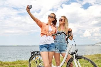 leisure, technology and friendship concept - happy smiling teenage girls or friends with bicycle taking selfie by smartphone at seaside in summer. teenage girls with bicycle taking selfie in summer