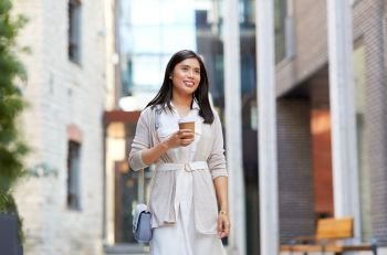 lifestyle and people concept - happy smiling young asian woman with takeaway coffee cup walking along city street. smiling woman with takeaway coffee cup in city