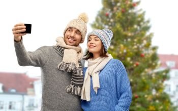 people, technology and winter holidays concept - happy couple in knitted hats and scarves taking selfie by smartphone over christmas tree in old town of tallinn city background. couple taking selfie at christmas tree in tallinn