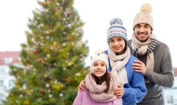 family and winter holidays concept - happy mother, father and little daughter in knitted hats and scarves over christmas tree in old town of tallinn city background. happy family over christmas tree in tallinn