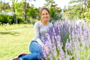 gardening and people concept - happy young woman sitting on grass near lavender flowers on summer garden bed. young woman and lavender flowers at summer garden