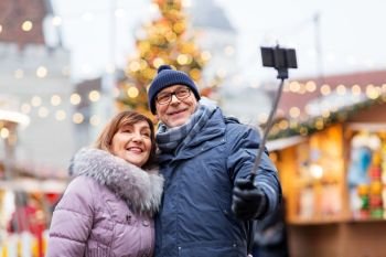 technology, winter holidays and people concept - happy senior couple taking picture by smartphone on selfie stick at christmas market on town hall square in tallinn, estonia. senior couple taking selfie at christmas market