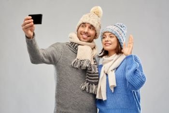 people, christmas and winter clothes concept - happy couple in knitted hats and scarves taking selfie by smartphone over grey background. couple in winter hats taking selfie by smartphone