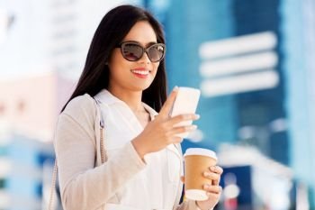 communication, lifestyle and technology concept - happy smiling young asian woman in sunglasses with takeaway coffee cup and smartphone on city street. smiling woman with smartphone and coffee in city