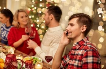 holidays, communication and celebration concept - happy young man calling on smartphone and having christmas dinner with friends at home. man calling on smartphone at christmas dinner