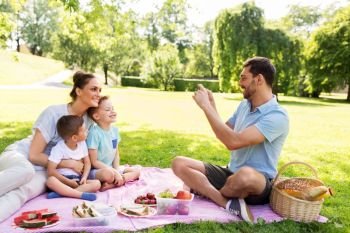 family, leisure and people concept - father taking picture of happy mother with two little sons by smartphone on picnic at summer park. father taking picture of family on picnic at park