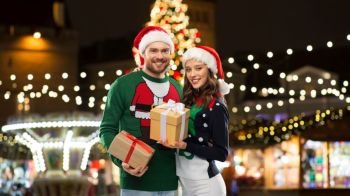 people and holidays concept - happy couple in santa hats and ugly sweaters with gifts over christmas market lights background. happy couple with gifts at christmas market
