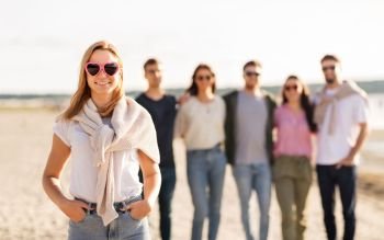 friendship, valentine’s day and people concept - happy woman in heart-shaped sunglasses with group of friends on beach in summer. happy woman with friends on beach in summer