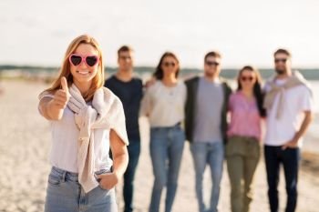 friendship, valentine’s day and people concept - happy woman in heart-shaped sunglasses with group of friends on beach in summer showing thumbs up. woman with friends on beach showing thumbs up