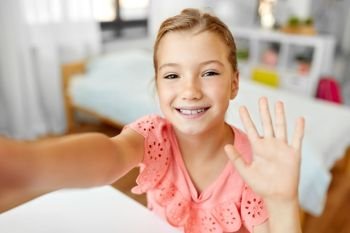 people, children and technology concept - girl taking selfie and waving hand at home. happy girl taking selfie and waving hand at home