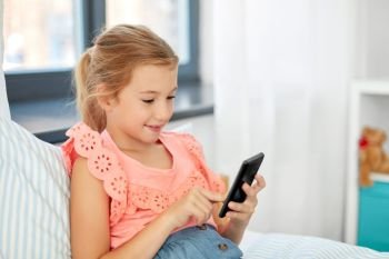 children, technology and communication concept - smiling girl using smartphone sitting on bed at home. girl using smartphone sitting on bed at home