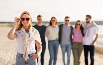 friendship, valentine’s day and people concept - happy woman in heart-shaped sunglasses with group of friends on beach in summer. happy woman with friends on beach in summer
