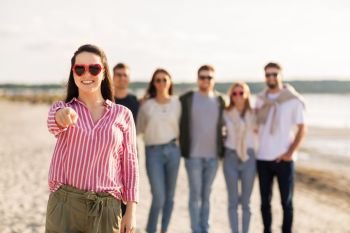 friendship, valentine’s day and people concept - happy woman in heart-shaped sunglasses with group of friends on beach in summer pointing to you. woman with friends on beach pointing to you