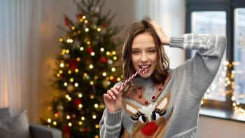 people and holidays concept - happy young woman wearing ugly sweater with reindeer pattern biting candy cane over home and christmas tree lights on background. woman in christmas sweater biting candy cane