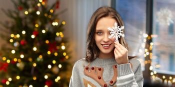 people and holidays concept - happy young woman with snowflake decoration wearing ugly sweater over home and christmas tree lights on background. woman in christmas sweater with snowflake at home