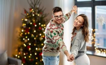 celebration, fun and holidays concept - happy couple wearing ugly sweaters dancing over christmas tree lights and home background. couple dancing in christmas ugly sweaters at home