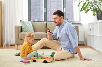 family, fatherhood and people concept - father with smartphone and little baby daughter playing with wooden toy toy blocks kit at home. father with smartphone and baby daughter at home