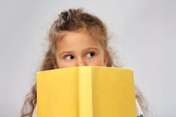 reading, education and childhood concept - little girl hiding behind yellow book over grey background. little girl hiding behind yellow book