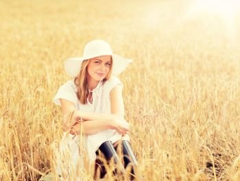 nature, summer holidays, vacation and people concept - happy young woman in white dress, rubber boots and sun hat sitting on cereal field. happy young woman in sun hat on cereal field