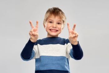 childhood, gesture and people concept - portrait of smiling little boy in striped pullover showing peace hand sign over grey background. little boy in striped pullover showing peace