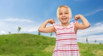 childhood and people concept - smiling little girl in striped dress showing her power over summer meadow and blue sky background. smiling little girl showing her power in summer