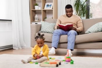 family, technology and people concept - happy african american father reading book and baby daughter playing toy blocks at home. happy father with tablet pc and baby at home