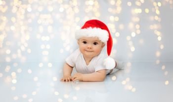 christmas, holidays and childhood concept - sweet little baby in santa helper hat lying on floor over festive lights background. little baby in santa helper hat lying on floor