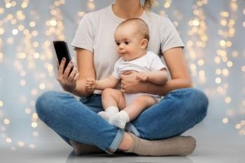 family, technology and motherhood concept - mother and baby looking at smartphone over festive lights background. mother and baby looking at smartphone over lights