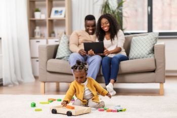childhood and people concept - little african american baby girl playing with toy blocks and parents with tablet pc computer at home. family with tablet pc and toy blocks at home