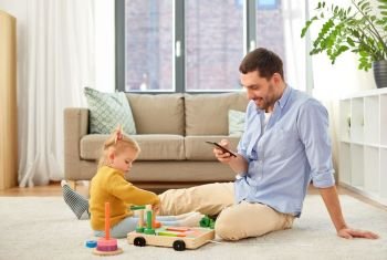 family, fatherhood and people concept - happy father with smartphone and little baby daughter playing with wooden toy toy blocks kit at home. father with smartphone and baby daughter at home