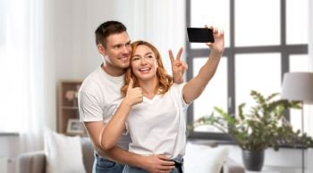 relationships, technology and people concept - happy couple in white t-shirts taking selfie smartphone and showing peace by over home background. happy couple in white t-shirts taking selfie