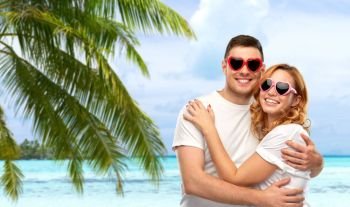 summer accessory, valentine’s day and love concept - portrait of happy couple in white t-shirts and heart shaped sunglasses over tropical beach background in french polynesia. happy couple in white t-shirts and sunglasses