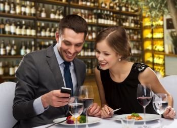 leisure and luxury concept - smiling couple with smartphone and food over restaurant or wine bar background. smiling couple eating main course at restaurant