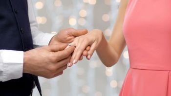 engagement, proposal and people concept - close up of man putting diamond ring on womans finger over holiday lights background. man puts engagement ring on womans finger