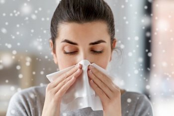 healthcare, cold, allergy and people concept - close up of sick woman blowing her runny nose in paper tissue at home in winter over snow. sick woman blowing nose in paper tissue at home