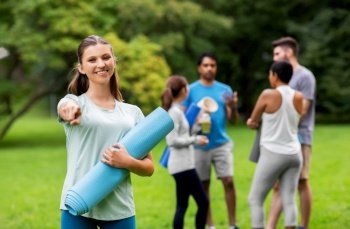 fitness, sport and healthy lifestyle concept - happy smiling young woman with mat pointing finger to camera over group of people meeting for yoga class at summer park. woman with yoga mat pointing finger to camera