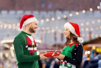 people and holidays concept - happy couple in santa hats and ugly sweaters with gift box over christmas market background. happy couple in christmas sweaters with gift box