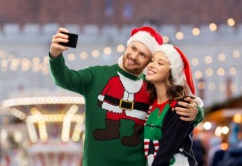 people, technology and holidays concept - happy couple in santa hats and ugly sweaters taking selfie with smartphone over christmas market background. happy couple in christmas sweaters taking selfie