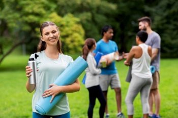 fitness, sport and healthy lifestyle concept - happy smiling young woman with mat and bottle over group of people meeting for yoga class at summer park. smiling woman with yoga mat and bottle at park