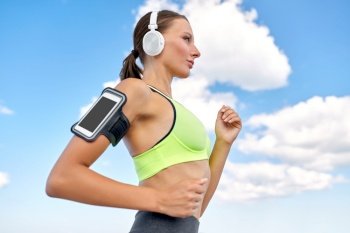 fitness, sport and healthy lifestyle concept - young woman with headphones and smartphone in armband running over blue sky. woman with headphones and smartphone running