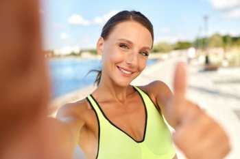 people, sport and fitness concept - portrait of smiling young sporty woman taking selfie picture and showing thumbs up outdoors on sea promenade. sporty woman taking selfie and showing thumbs up