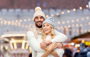 winter holidays and people concept - smiling man and woman in hats and scarf hugging over christmas market background. couple in winter hugging over christmas market
