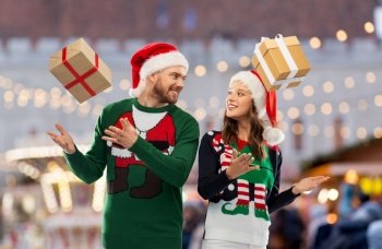 people and holidays concept - happy couple in santa hats and ugly sweaters with gifts over christmas market background. happy couple in christmas sweaters with gifts