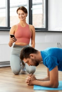 sport, fitness, lifestyle and people concept - smiling young woman with smartphone and man exercising and doing plank at home. happy couple exercising at home