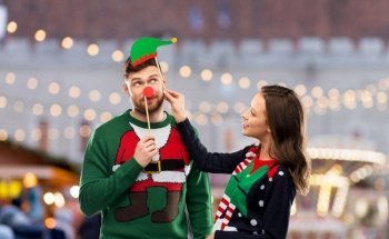 people and holidays concept - happy couple in ugly sweaters posing with party props over christmas market background. couple with christmas party props in ugly sweaters