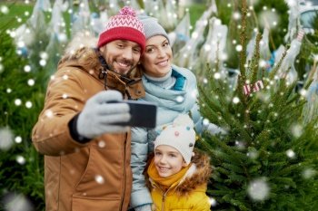 family, winter holidays and people concept - happy mother, father and little daughter buying christmas tree and taking selfie with smartphone at street market over snow. family taking selfie with christmas tree at market